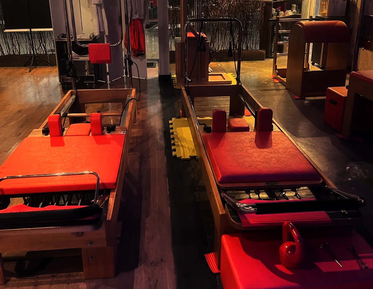Dois aparelhos de pilates de madeira clara e estofado vermelho, lado a lado, formando um corredor até a vitrine de galhos secos da entrada