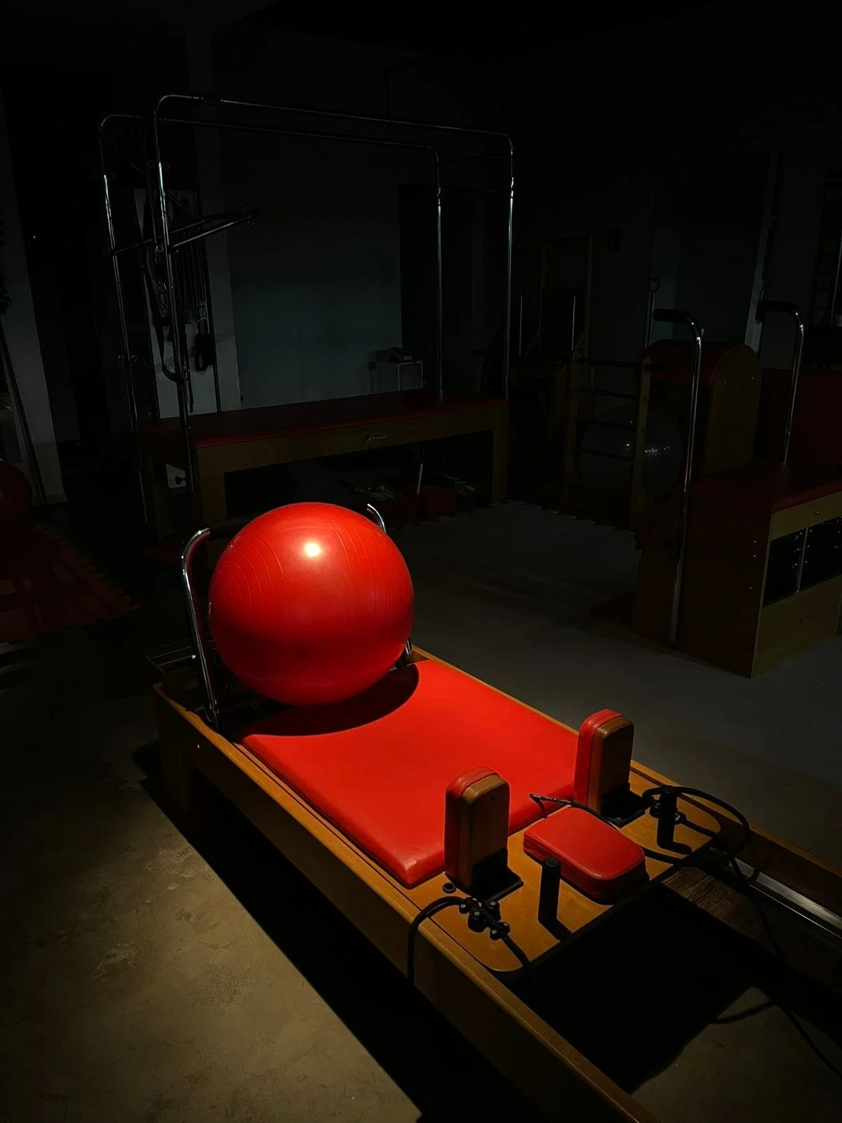 Bola vermelha sobre um aparelho de pilates de estofado vermelho, no salão da Clínica Rennova com as luzes baixas